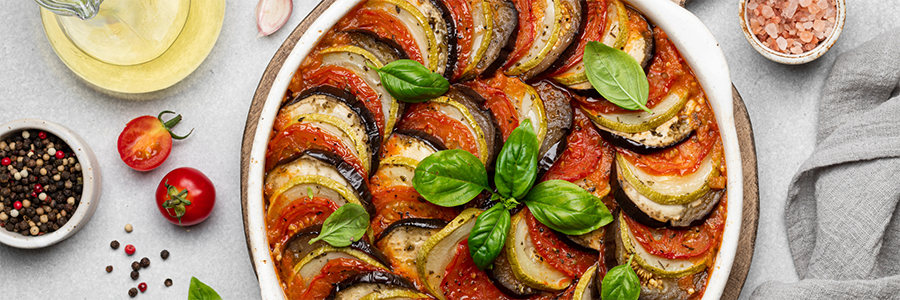 Wide shot of a round platter with ratatouille. Layers of sliced zucchini, squash, eggplant, and tomato and garnished with basil leaves.