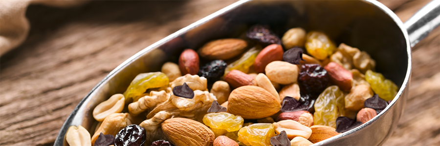 Close-up shot of a metal scoop filled with trail mix containing an assortment of nuts, dried fruit, and chocolate chips