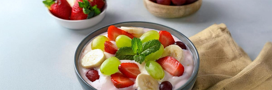 Wide shot of a bowl of fruit salad with green grapes, strawberries, and sliced bananas in strawberry yogurt. Salad is garnished with a sprig of mint. A small bowl of strawberries is displayed in the background but is blurred.