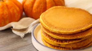 Shot of a stack of pancakes on a plate with small pumpkins displayed in the background.