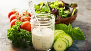 Jar of homemade ranch dressing with fresh vegetables displayed next to it on a table.