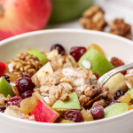 Wide shot of a bowl of oatmeal topped with chopped apples, raisins and nuts. Raisins are displayed around the bowl as presentation.
