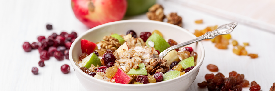 Wide shot of a bowl of oatmeal topped with chopped apples, raisins and nuts. Raisins are displayed around the bowl as presentation.