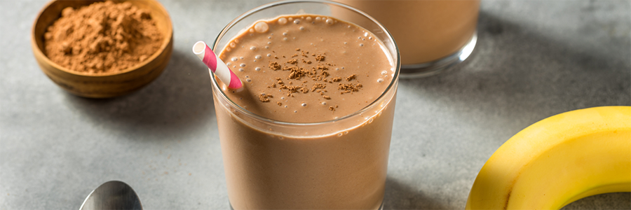 Wide shot of a table with a clear glass with a chocolate banana shake and straw. There is a ripe banana and small bowl of cocoa displayed nearby.