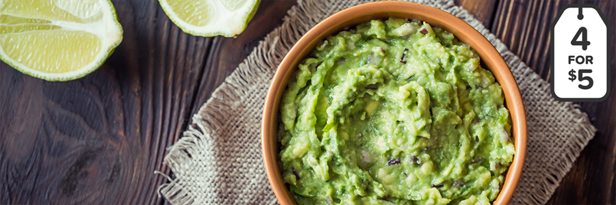 Wide shot of a wood table with a bowl of guacamole in a stoneware bowl. Bowl is resting on a linen napkin and table is displayed with limes cut in half.