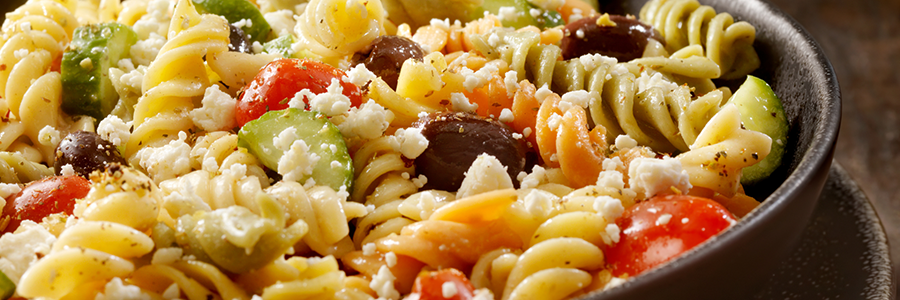 Close up of a bowl with rotini pasta, tomatoes, cucumbers, olives, and feta cheese.
