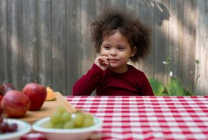 A small child at a table with gingham table cloth. The table is det with fruit like apples, melon, and grapes. The child is looking at the fruit, contemplating.