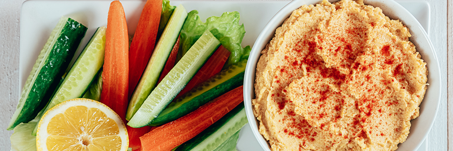 Close up shot of a bowl of chickpea dip garnished with paprika. A platter with sliced carrots, peppers, and cucumbers is displayed next to it.