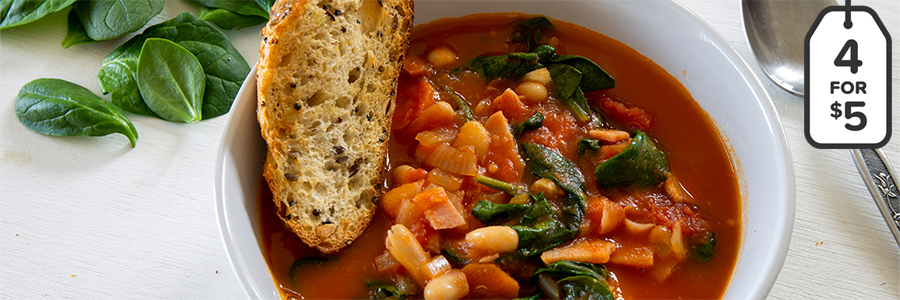 Close up of tomato based soup with white beans, tomatoes, and spinach in a white bowl. Slice of toasted bread on the side of the bowl with spinach leaves and silver spoon.