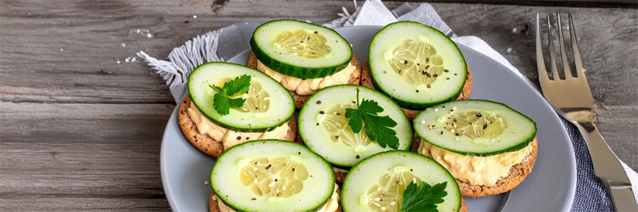 A plate with crackers topped with hummus and sliced cucumbers.