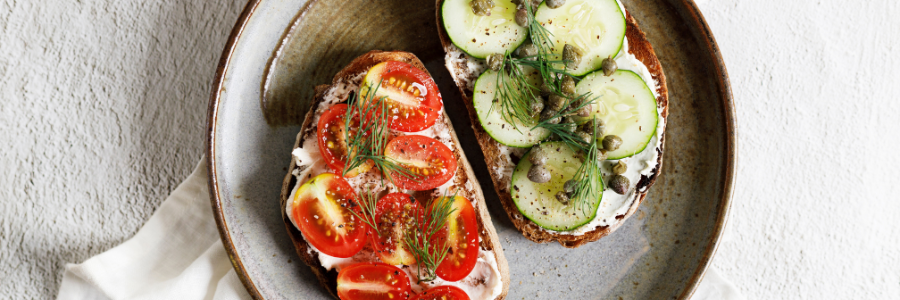 Overhead shot of a plate with two slices of toast, one topped with cheese spread and grape tomatoes, the other topped with cheese spread, sliced cucumbers and capers.