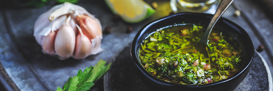 Small bowl of chimichurri sauce of a dining table. Table is also displayed with herbs, lime wedges, and a bulb of garlic.