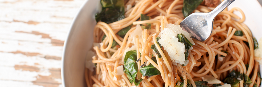 Close up shot of a white bowl filled with a mix of spaghetti, kale, and shaved Parmesan cheese.