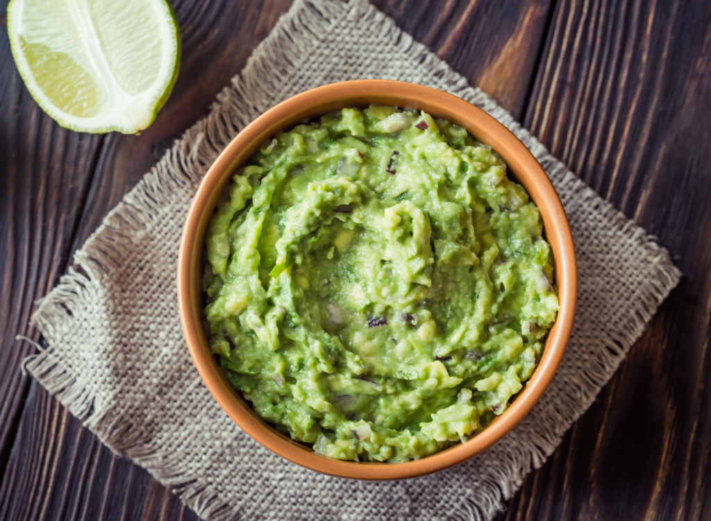 Shot of a wood table with a bowl of guacamole in a stoneware bowl. Bowl is resting on a linen napkin and table is displayed with limes cut in half.