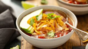 Bowl of tortilla soup with toppings like avocado slices, tortilla strips and cilantro.